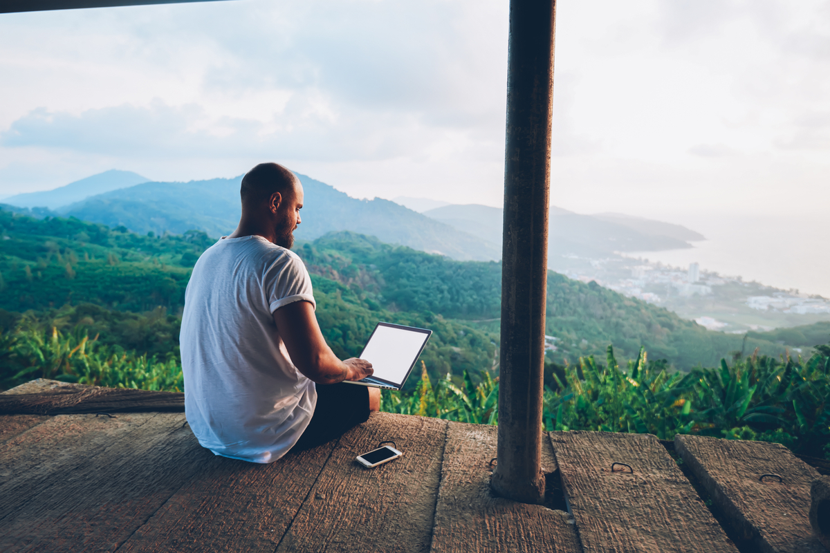 Jovem sentado em uma varanda em meio à natureza, utilizando seu notebook para trabalhar remotamente