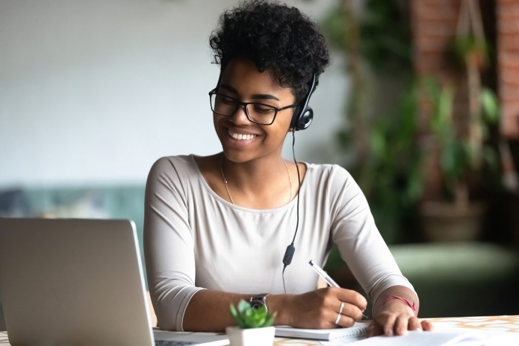 Mulher jovem, sorridente, com fones de ouvido, anotando algo em um caderno enquanto observa a tela do seu notebook