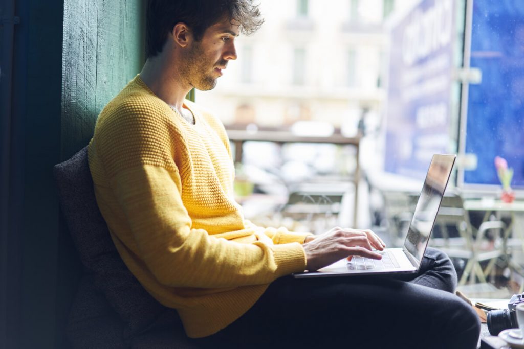 Jovem com um notebook no colo em uma espécie de cafeteria