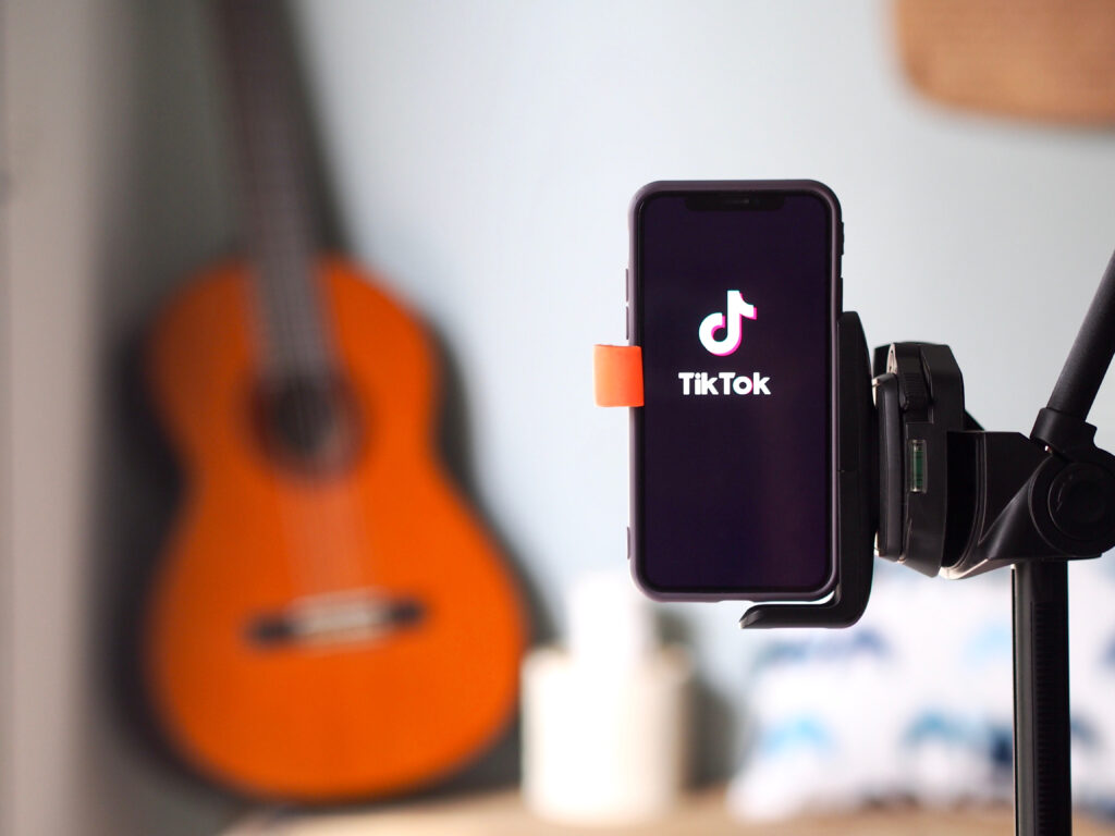 Imagem de um celular acessando o TikTok, em primeiro plano, preso em um pedestal, com um violão ao fundo, em segundo plano