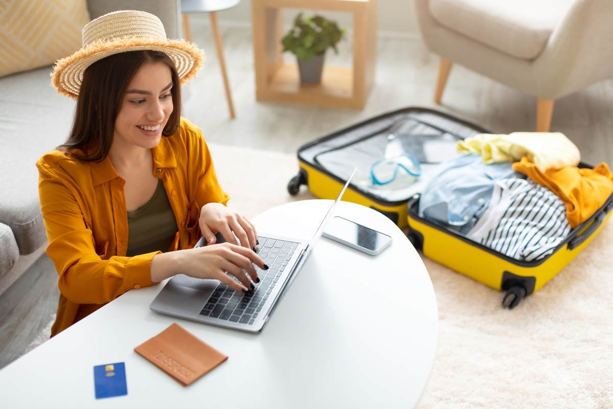 Mulher pesquisando como vender milhas no notebook com mala de viagem aberta ao lado.