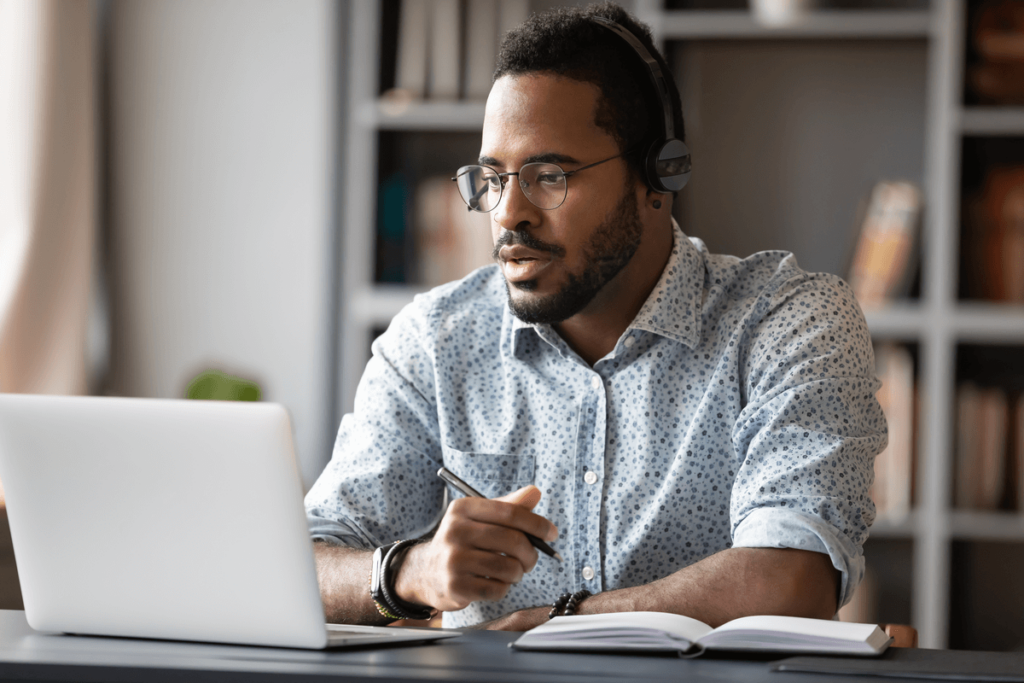 Homem usando o notebook segurando uma caneta e usando um caderno ao lado