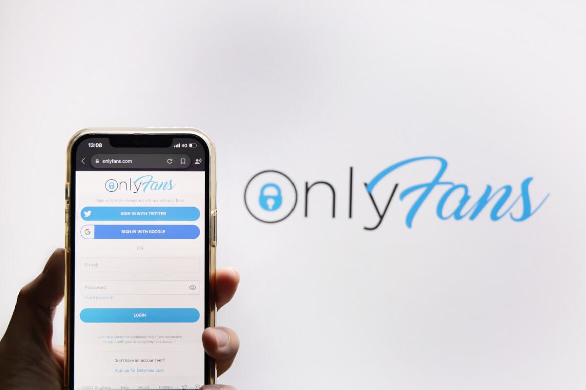 Uma parede com o logo do OnlyFans e em frente uma mão com um celular abrindo o app do OnlyFans