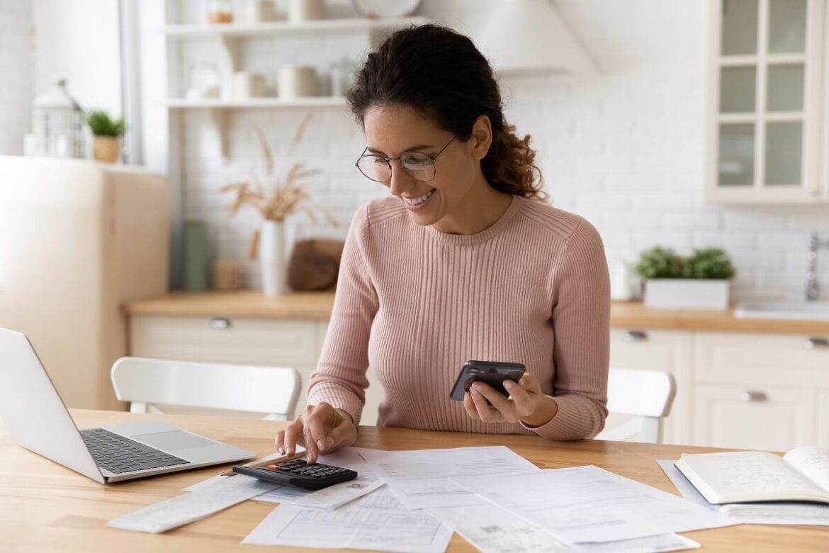 Mulher em uma mesa com notebook, documentos e usando uma calculadora e o celular