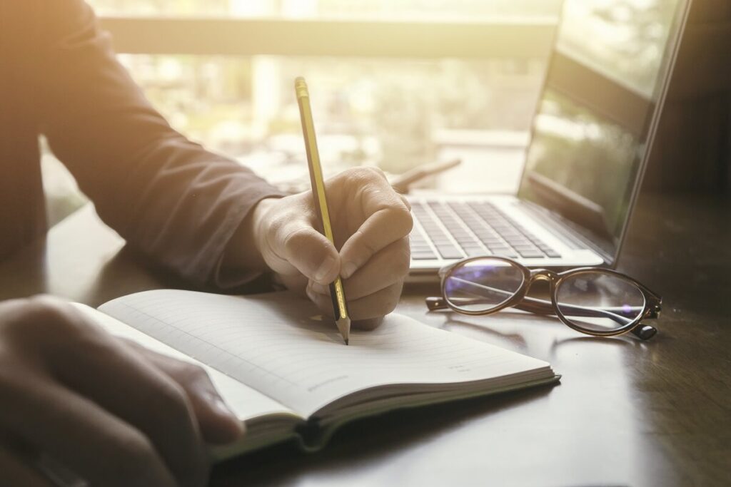 Pessoa produzindo conteúdo escrevendo em um caderno com o computador aberto do lado