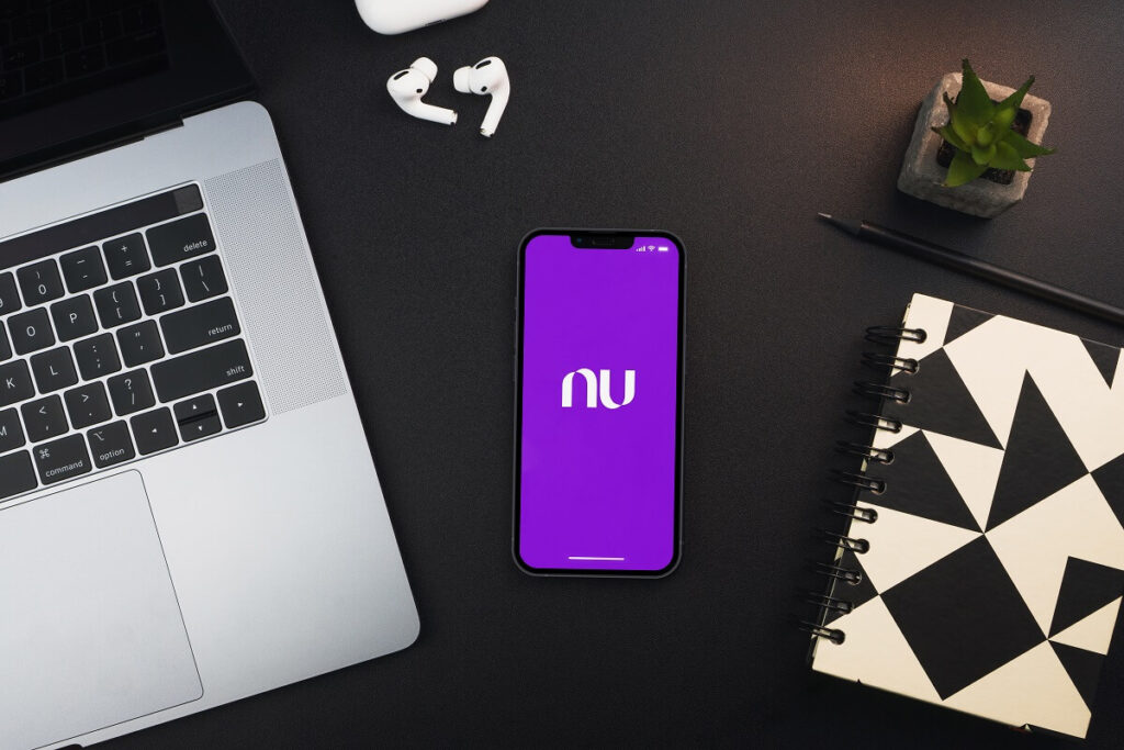 Celular com o app Nubank aberto e em volta um notebook aberto, um caderno e um fone sem fio lado