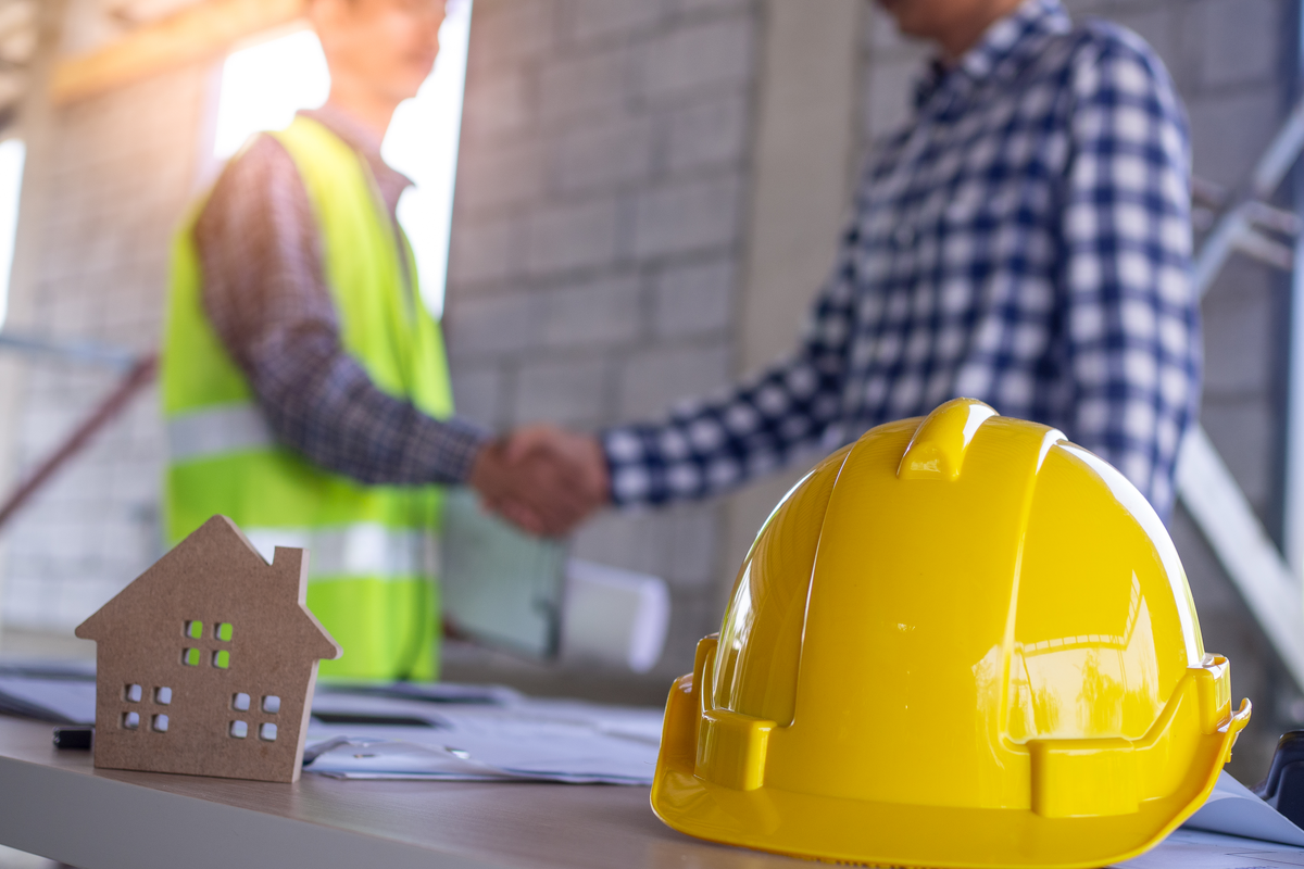 Em primeiro plano, um capacete de construção e uma placa de MDF em formato de casa, em segundo plano, dois homens apertando as mãos