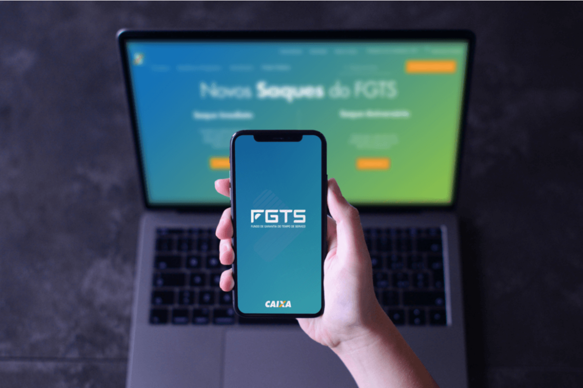 Computador com a tela do saque do novo FGTS em segundo plano, em primeiro plano uma mão segurando um celular com o aplicativo do FGTS aberto