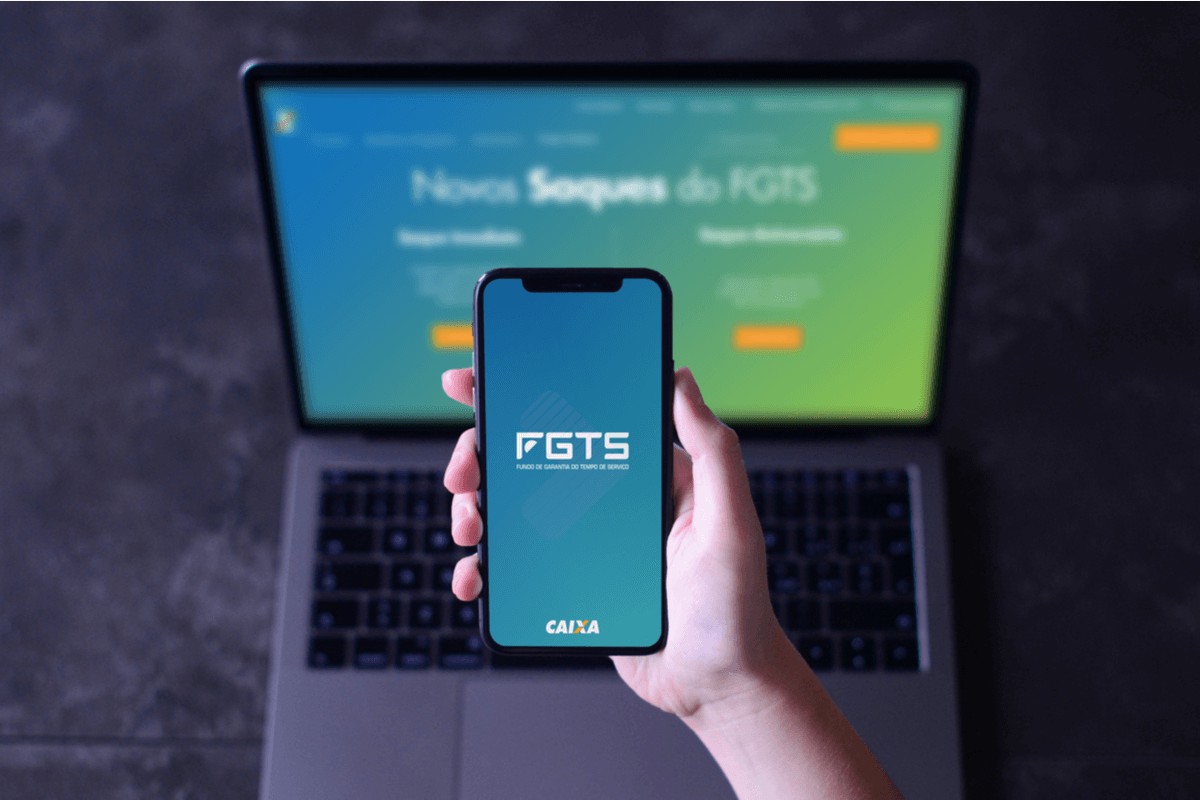 Computador com a tela do saque do novo FGTS em segundo plano, em primeiro plano uma mão segurando um celular com o aplicativo do FGTS aberto