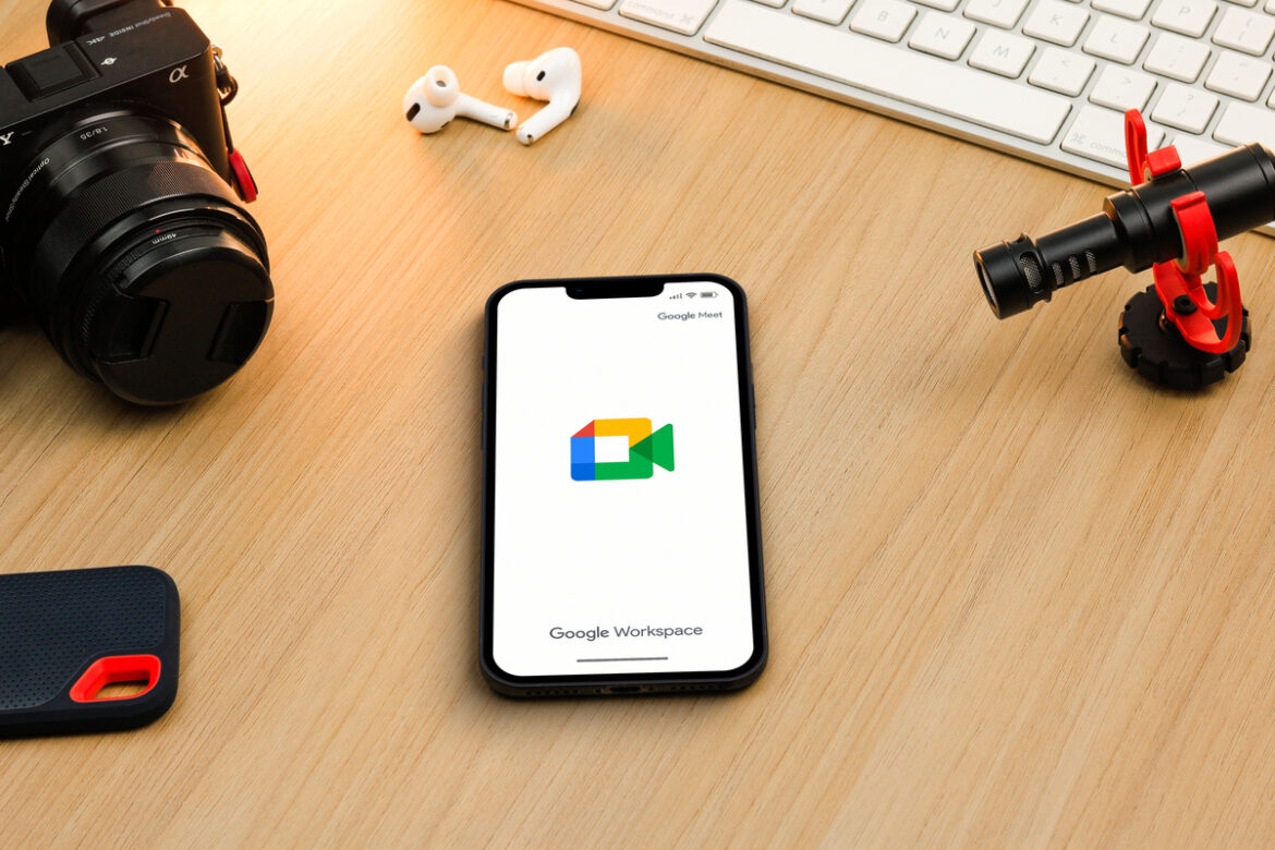 Um celular no cento de uma mesa com o logo do Google Meet, cercado por itens de criadores de conteúdo, como câmera, fones de ouvido e computador.
