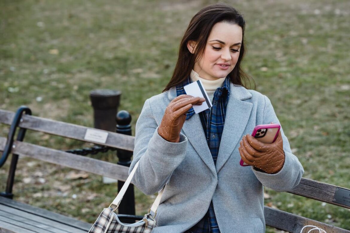 Uma mulher está de pé segurando um celular com a mão direita e com a mão esquerda segurando um cartão de crédito internacional