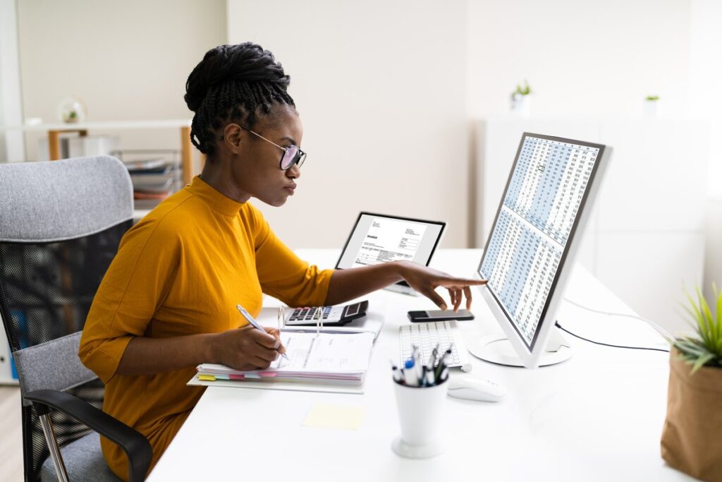 Mulher revisando os números na tela do computador e anotando na prancheta.