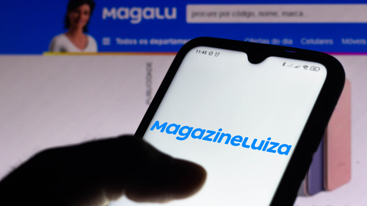 Imagem de uma pessoa segurando um celular. Na tela do celular, está carregando uma página da Magazine Luiza. Ao fundo, podemos ver um computador que também está conectado a uma página da Magazine Luiza.