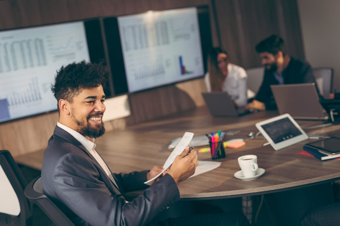 conomistas em uma sala de reunião discutindo gráficos e dados financeiros em celebração ao Dia do Economista, com um profissional sorridente segurando documentos em primeiro plano.